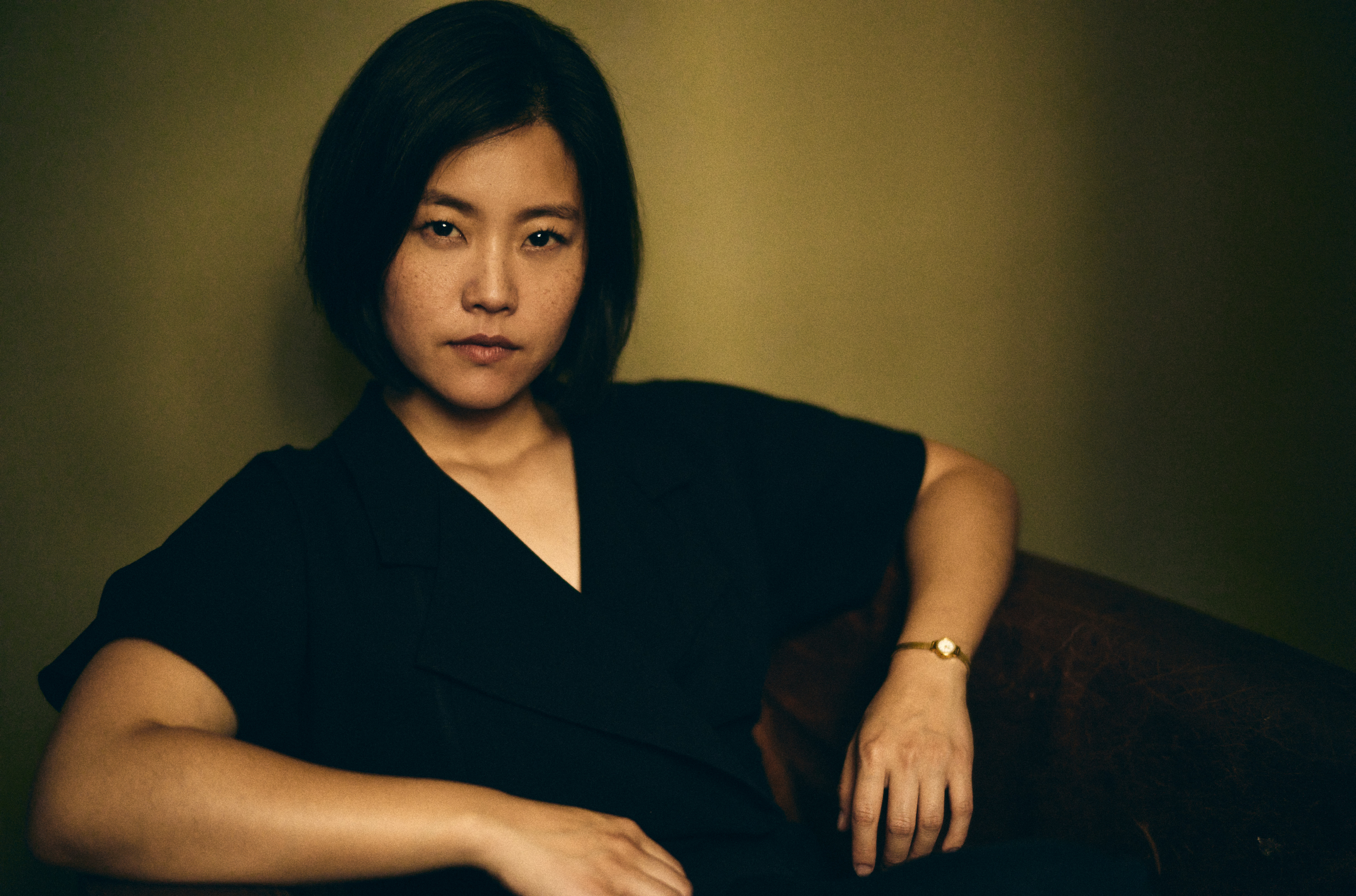 Mid-shot of a young East Asian woman reclining in an armchair in front of an olive-green background.