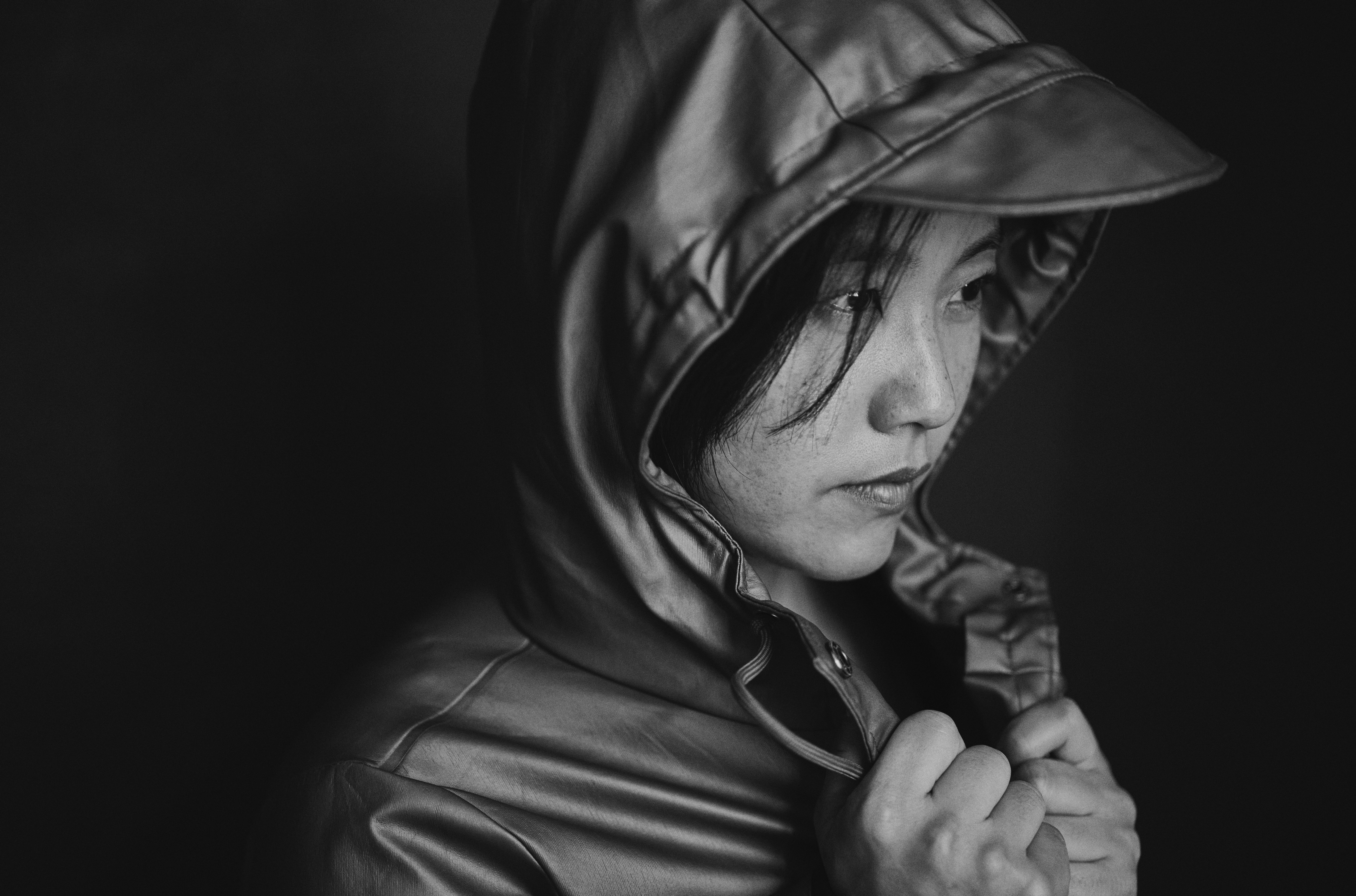 Monochrome mid-shot of an East Asian woman with freckles with the hood up and pulling on the lapel of her coat.