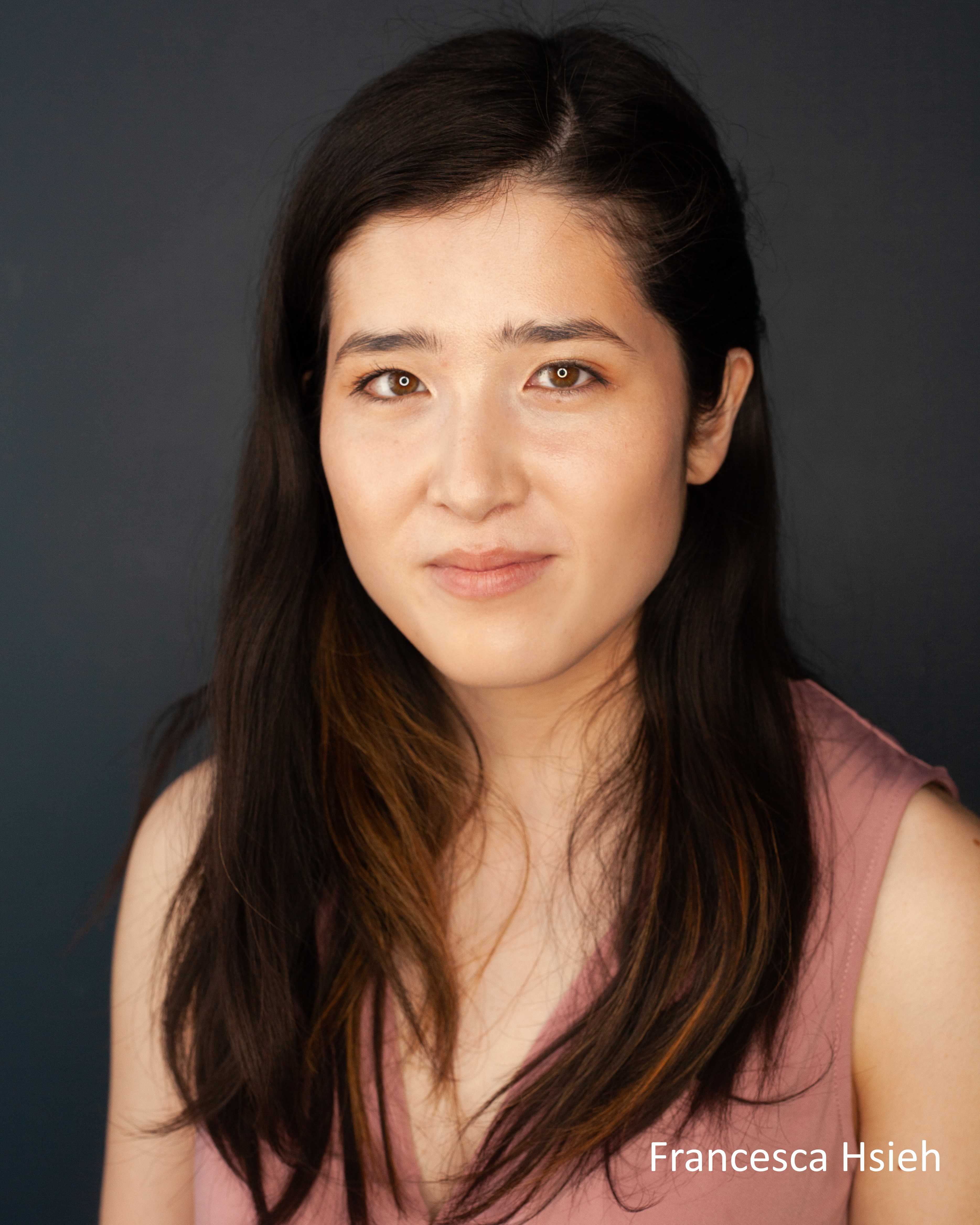 A headshot of a young mixed race East Asian woman with long hair down her pink top, smiling a little to camera.