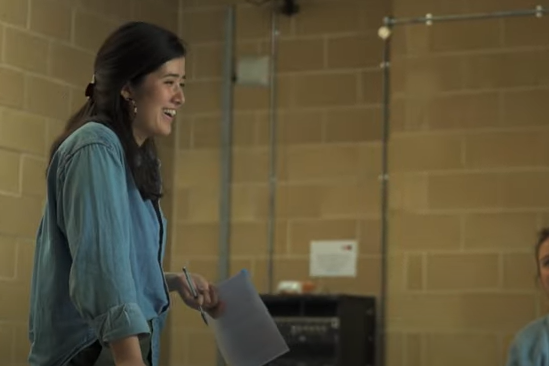 A young mixed race East Asian woman’s profile on the left-hand-side, holding a script and laughing in a brown-brick-walled rehearsal space.