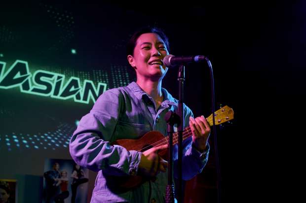 A young East-Asian woman is playing the ukulele and singing into the microphone in front of a green-glowing projection of "ASIAN"