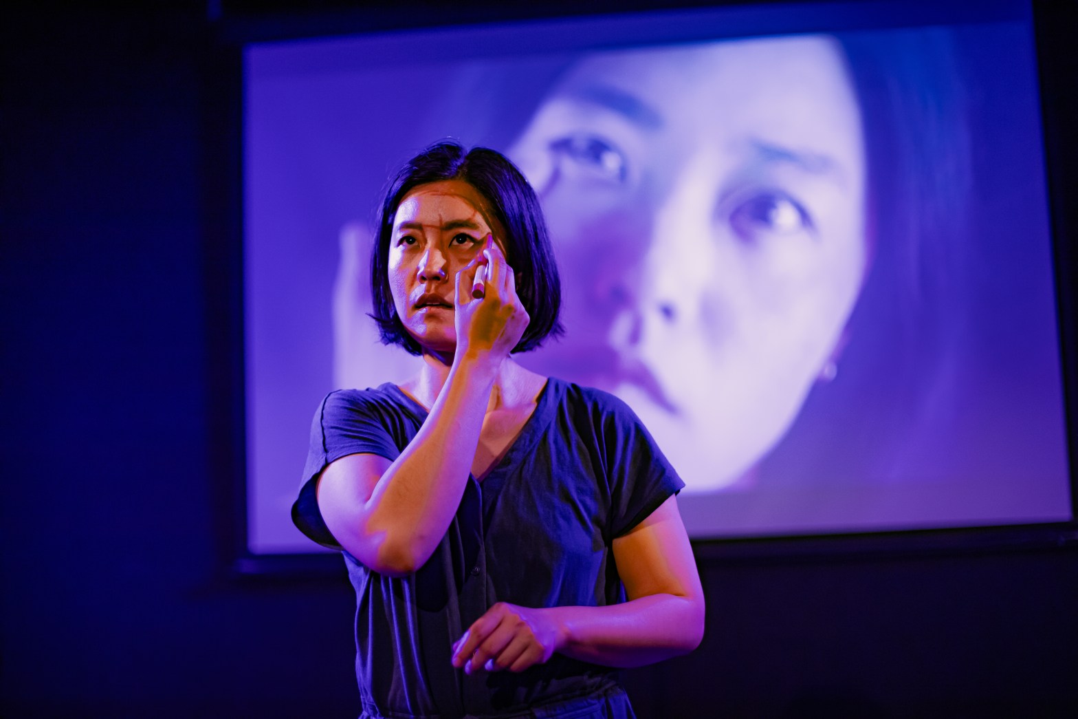 A young East Asian woman is drawing oh her own face with a lipstick in front of a video projection of her face close up, scrutinising her own face.